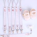 A set of baby accessories laid out on a white background, including five white leather suspenders with metal clips, a matching long strap, a pair of cream crocheted baby shoes, and a pacifier clip.