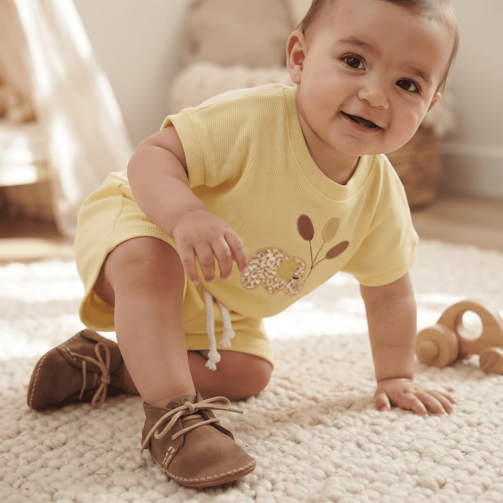 A baby crawling on a rug wearing Safari Vellies full leather grain shoes and a yellow outfit, smiling happily.
