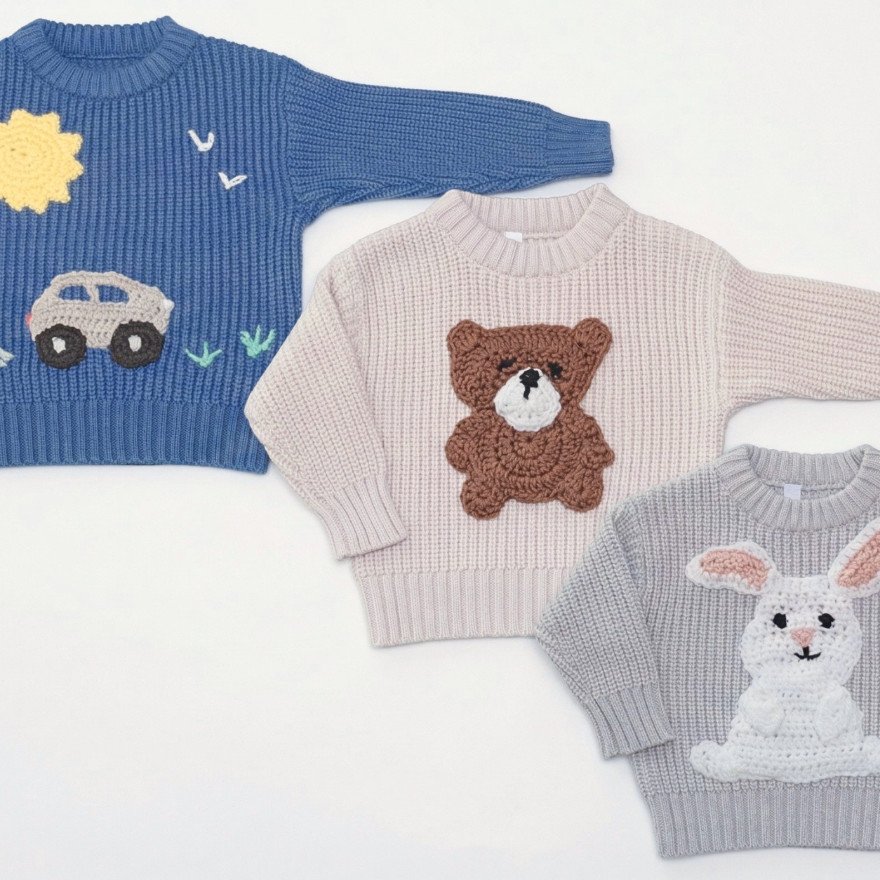 Three children's knit sweaters laid out on a white background: a blue sweater with a yellow sun and car motif, a pale pink sweater with a brown bear motif, and a light gray sweater with a white rabbit motif.