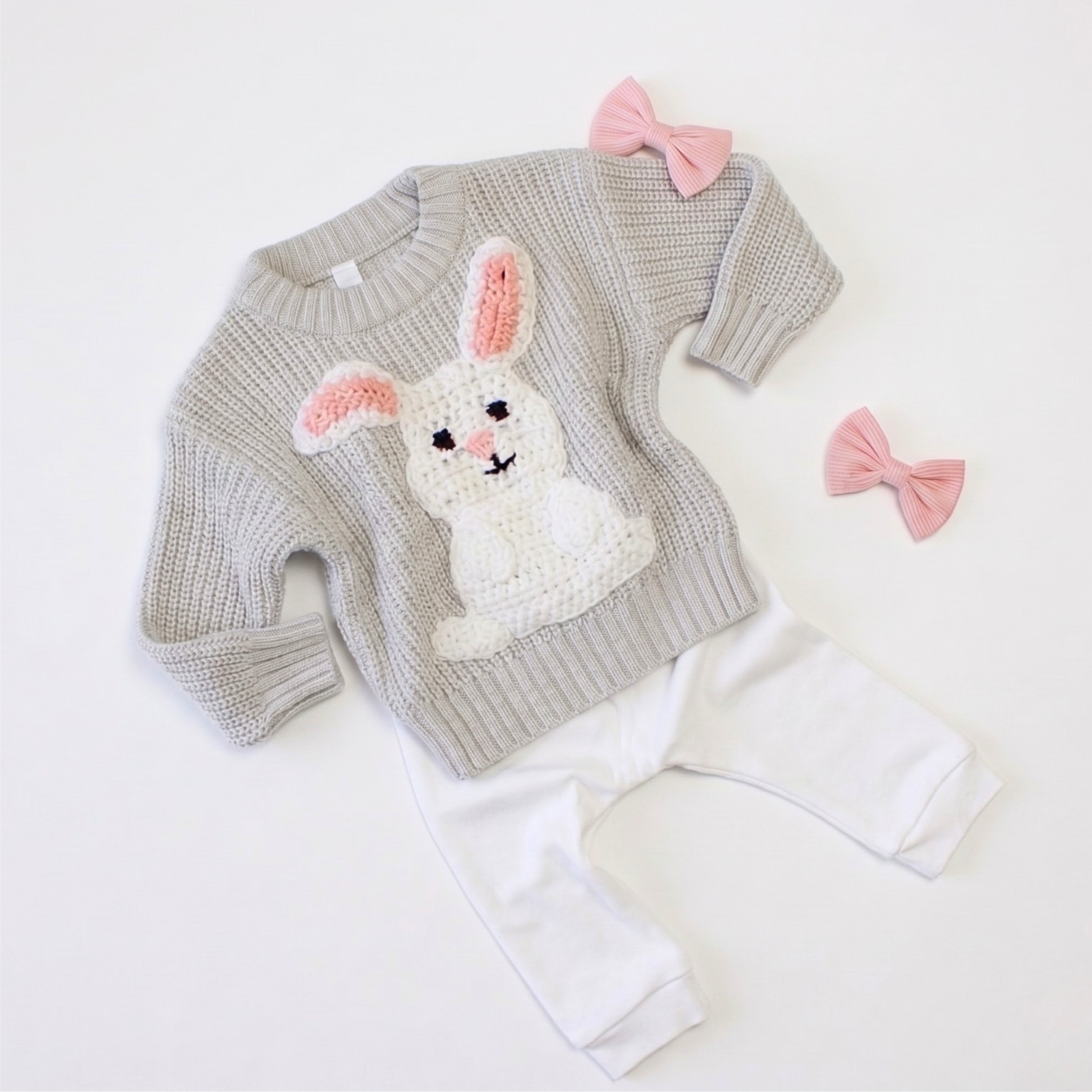 A baby clothing set featuring a light gray ribbed knit sweater with a white crocheted bunny applique on the front, paired with white pants. The set is styled with two pink fabric bows on a clean white background.