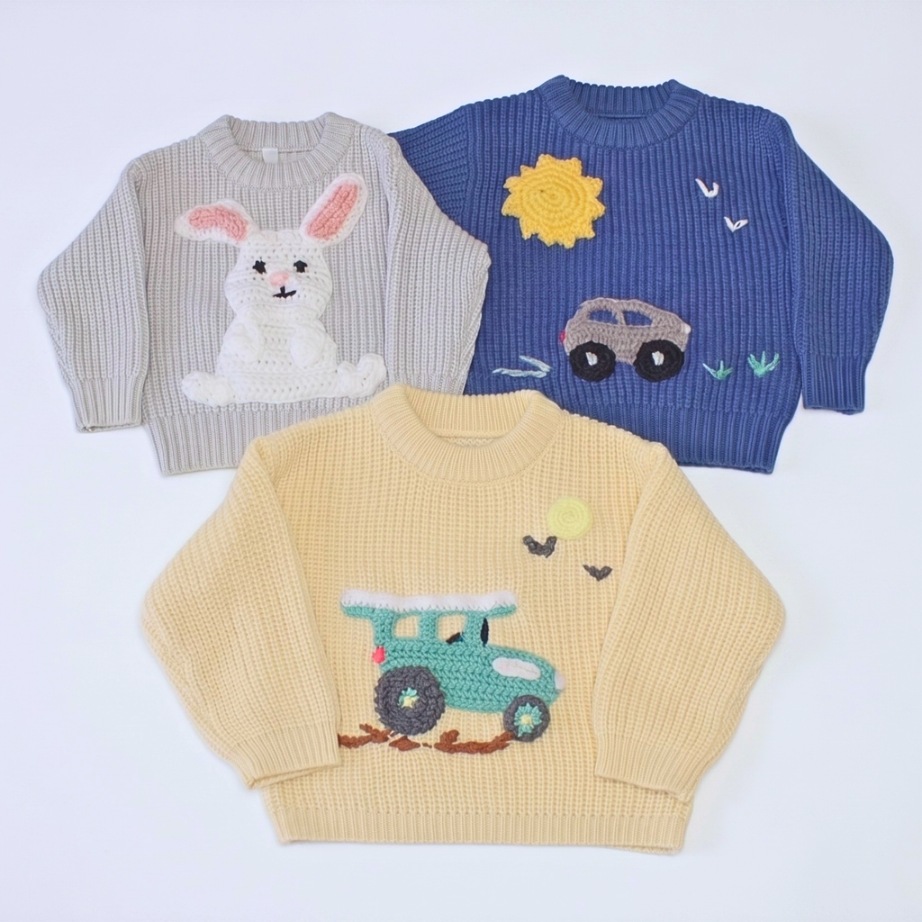 Three children's ribbed knit sweaters laid flat on a white background: a light gray sweater with a white bunny motif, a royal blue sweater with a yellow sun and car motif, and a pale yellow sweater with a teal truck motif.